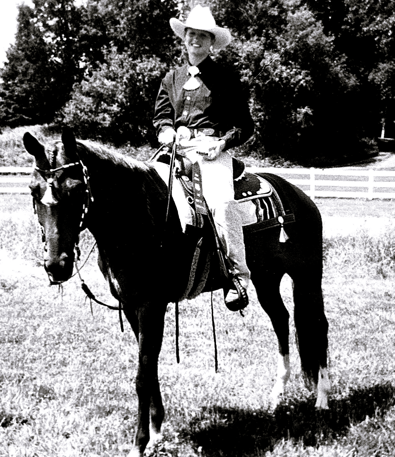 Beth Crain as a child, in western show clothes and a white hat, seated on a dark horse with a white blaze in a grass paddock.